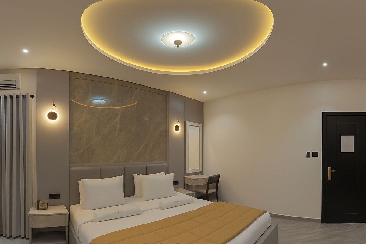 A modern hotel room with a large bed, a round ceiling light, a mirror, and a door.