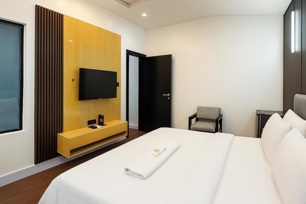 A modern hotel room with a large bed, a TV mounted on a wooden panel, a small armchair, and a bedside table.