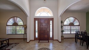 Un hall d’entrée spacieux avec une grande porte en bois, deux fenêtres en arc surmontées de stores, un sol carrelé et une petite table ornée d’un vase de fleurs.