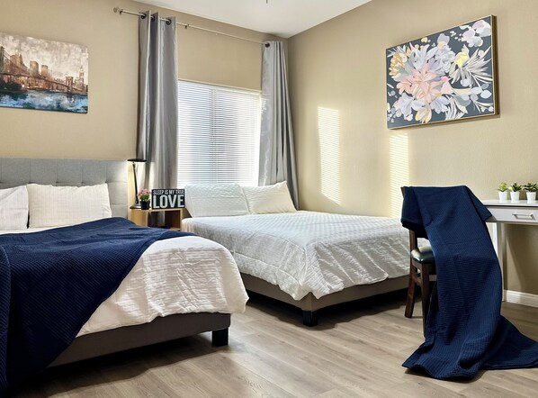A hotel room with two beds, a desk, a chair, and a view of a cityscape through the window.