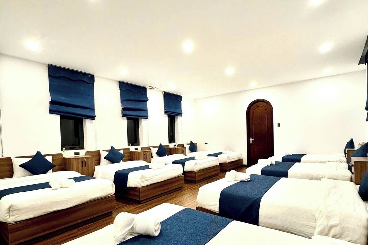 Habitación de hotel con varias camas dispuestas en fila, cada una con cabecero de madera y ropa de cama azul.