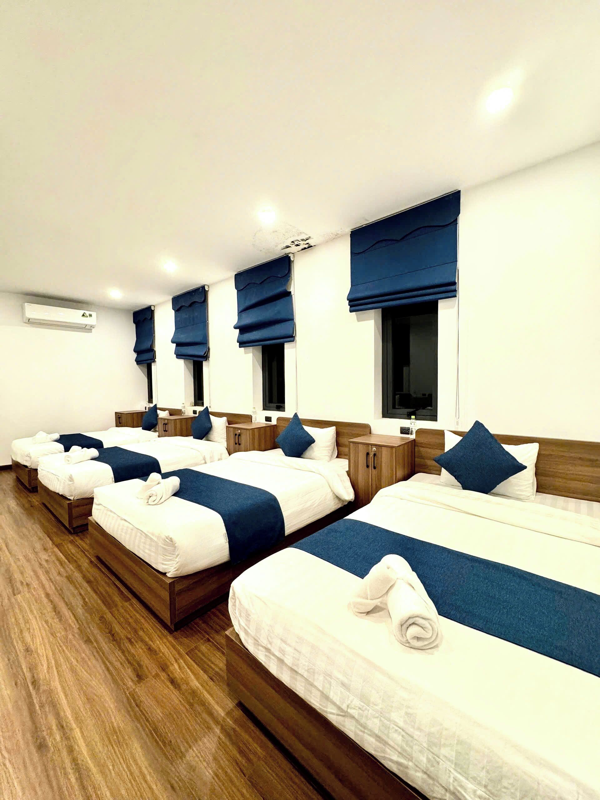 Ein Hotelzimmer mit vier Betten, die nebeneinander stehen, jedes mit blauer Bettdecke und weißen Laken. Jedes Bett hat ein Holz-Kopfteil, und der Boden des Zimmers ist aus Holz.