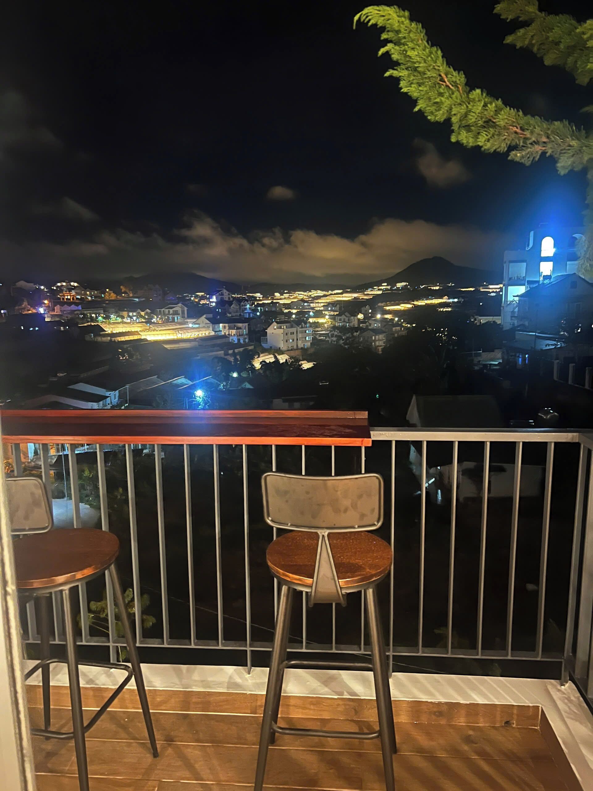A balcony with two metal chairs and a wooden table, overlooking a cityscape at night with illuminated buildings and a mountain in the background.