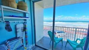 A balcony with a view of the beach, a chair, a table, and shelves with baskets and a laundry basket.