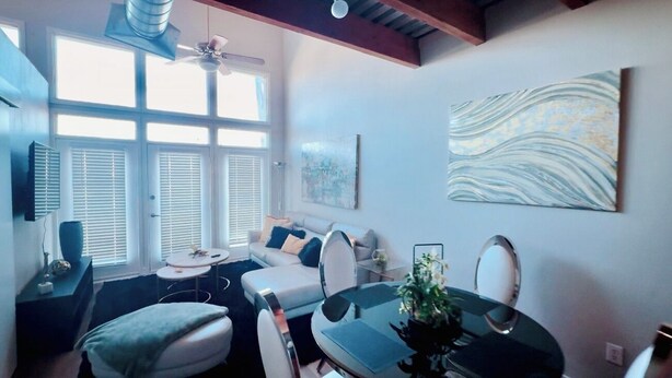 A modern living room with a sofa, dining table, and chairs. There is a large window with blinds, a ceiling fan, and artwork on the wall.