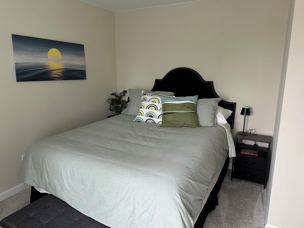 A neatly made bed with a grey bedspread and pillows, a black headboard, a wall-mounted painting of a sunset, a bedside table with a lamp, and a small plant. - Charming 6-bedroom house in marvelous Rochester Hills with a fitness room, WiFi (Rochester Hills)