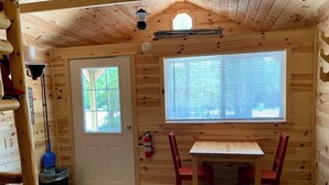 Interior de una cabaña con detalles de madera, una puerta, un ventanal, una mesa con sillas y un extintor.