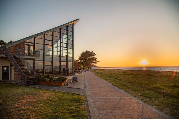 A modern building with large glass windows, a staircase, and a pathway leading to the beach.
