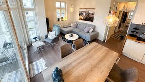 A modern living room with a grey sofa, a round coffee table, and a wooden dining table. There is a chair with a fluffy throw, a small side table, and a wall-mounted light fixture. - Vacation apartment Meehr Glowe - Villa Rosa - Holiday apartment Mee(h)r (Glowe)