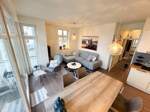 A modern living room with a grey sofa, a round coffee table, and a wooden dining table. There is a chair with a fluffy throw, a small side table, and a wall-mounted light fixture.