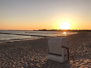 A beach with a white wicker chair in the foreground and a sunset in the background.