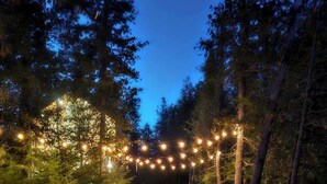 Un hogar con dos sillas Adirondack en un área boscosa al anochecer, rodeado de luces colgantes.