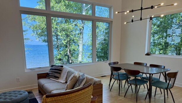Amplia sala de estar con un ventanal que da a un lago, comedor con mesa redonda y sillas, y una zona de estar acogedora con sofá y reposapiés.