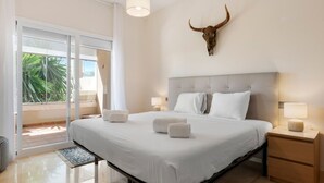A hotel room with a large bed, a nightstand with a lamp, a ceiling-mounted bull skull decoration, and a sliding glass door leading to a balcony with a view of palm trees. - Sunny Terrace Apt in Golf Valley with Pool (Estepona)