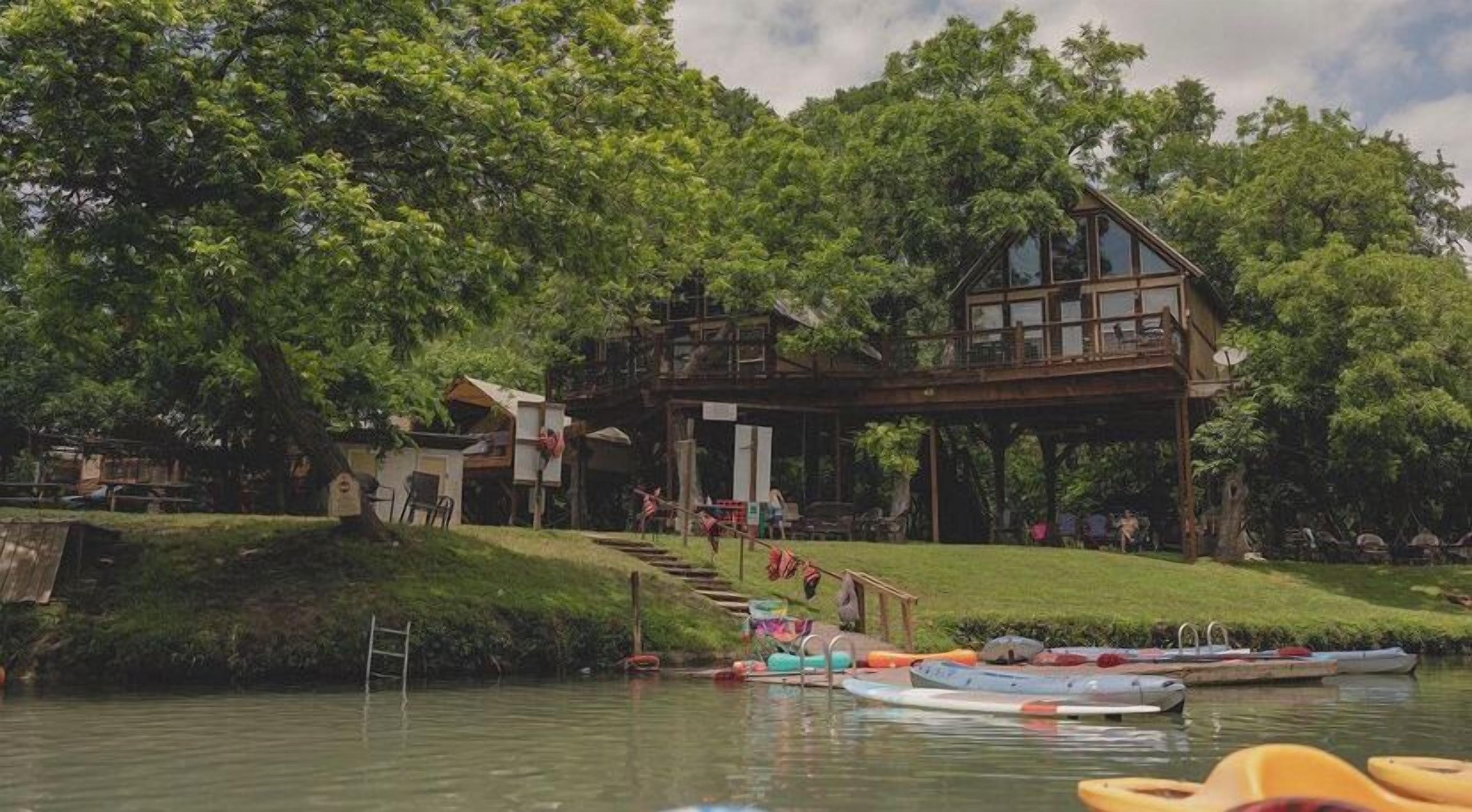 Una cabaña junto al lago con una terraza de madera, un espacio al aire libre cubierto y varias embarcaciones.