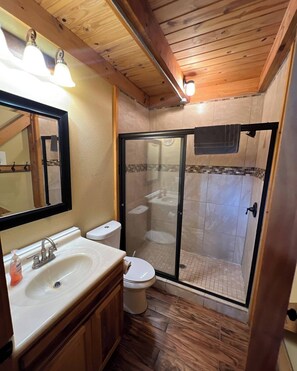 A bathroom with a wooden ceiling, a sink with a mirror, a toilet, and a glass-enclosed shower area. - Geronimo Creek Retreat (Seguin)