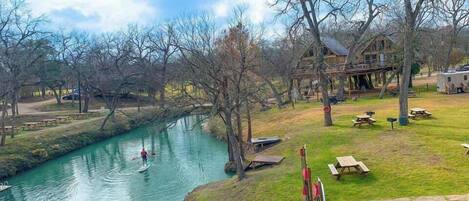A riverside view with a wooden dock, paddle boats, picnic tables, and a large house with a deck.