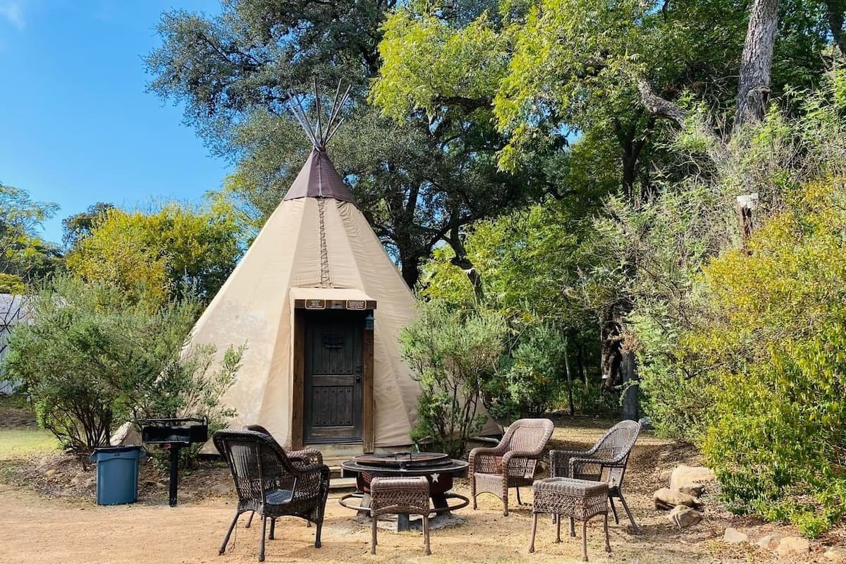 A conical tent with a door, surrounded by wicker chairs and a small table, set in a natural outdoor area.