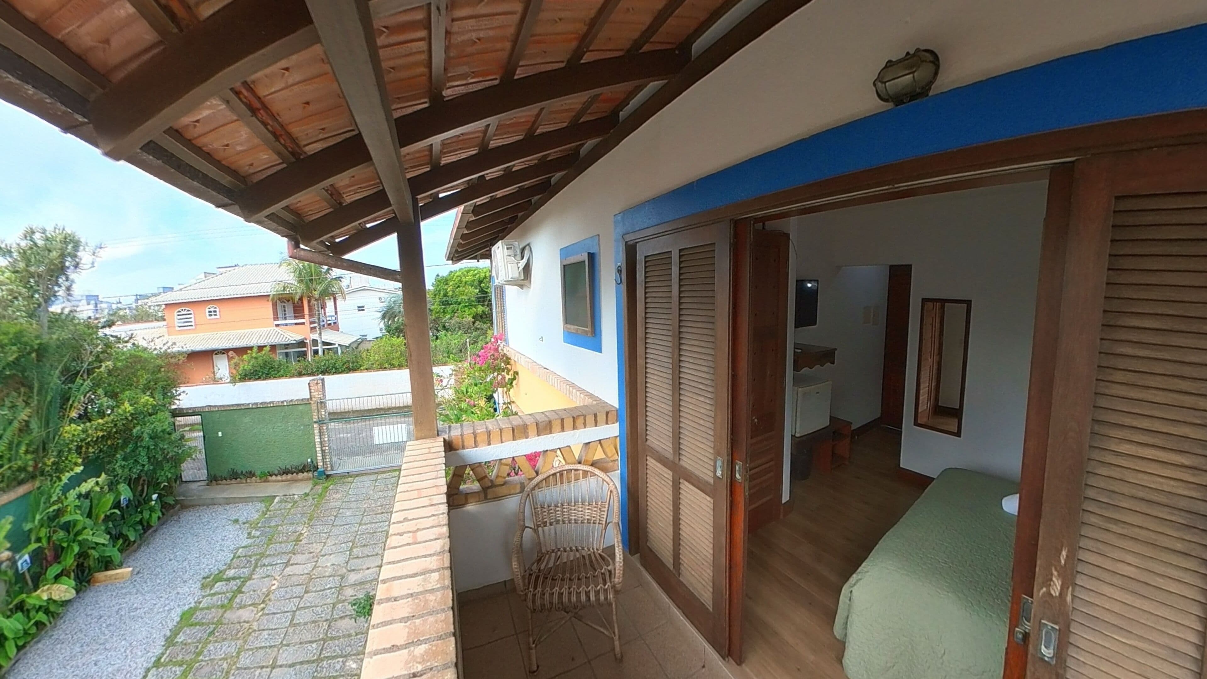 Un espacio al aire libre techado, con techo de madera, una silla de mimbre y un portón corredizo que da a una habitación con cama.