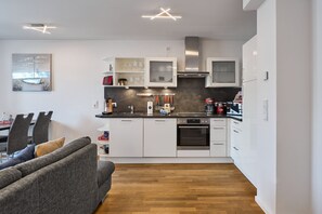 A modern kitchen with white cabinets, a built-in oven, and a dining area with a grey sofa and wooden flooring.