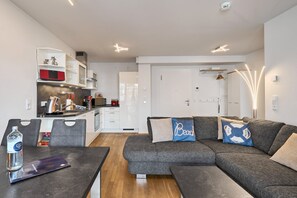 A modern living room with a grey sectional sofa, a dining table with chairs, and a kitchen area with white cabinets and a red appliance. - Moin by the sea! Hansastr. 5 a - Moin am Meer! Hansastr. 5 a 100 meters to the (Scharbeutz)