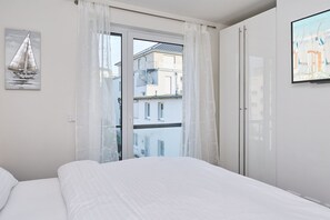 A bedroom with a bed, a window with sheer curtains, a painting of a sailboat, and a view of buildings outside. - Moin by the sea! Hansastr. 5 a - Moin am Meer! Hansastr. 5 a 100 meters to the (Scharbeutz)