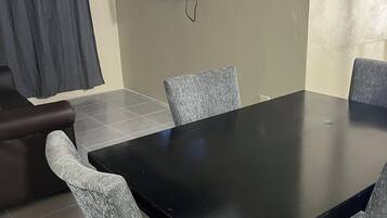 A dining area with a black table and four grey chairs, a television mounted on the wall, and a dark curtain on the left.