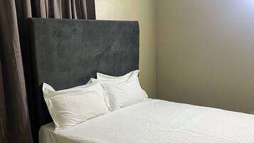 Un lit simple avec une tête de lit sombre et du linge de lit blanc, dans une pièce aux murs unis et au sol carrelé.