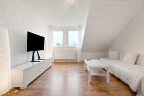 A modern, white-themed room with a TV, a white sofa, a small table, and a window with a view.