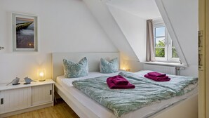 Un dormitorio con techo inclinado, cama con colcha estampada de flores verdes, dos almohadas, mesita de noche con lámpara y un cuadro en la pared.