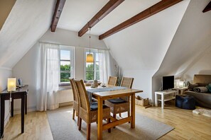 Een knus zolderkamer met een houten eettafel, rieten stoelen, een bank en een televisie.