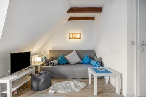 Ein modernes Wohnzimmer mit Flachbildfernseher, einem grauen Sofa mit blau-weißen Kissen, einem weißen Couchtisch, einem kleinen weißen Teppich und einer Holzbalkendecke.