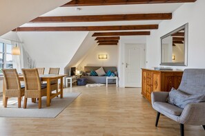 A loft-style living area with exposed wooden beams, a dining table with chairs, a sofa, a gray armchair, and a wooden cabinet.