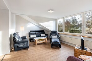 A modern living room with a sofa, two armchairs, a coffee table, and a TV on a wooden box. Large windows provide natural light and a view of the outside. - Holiday flat in Schulensee near Kiel (Molfsee)
