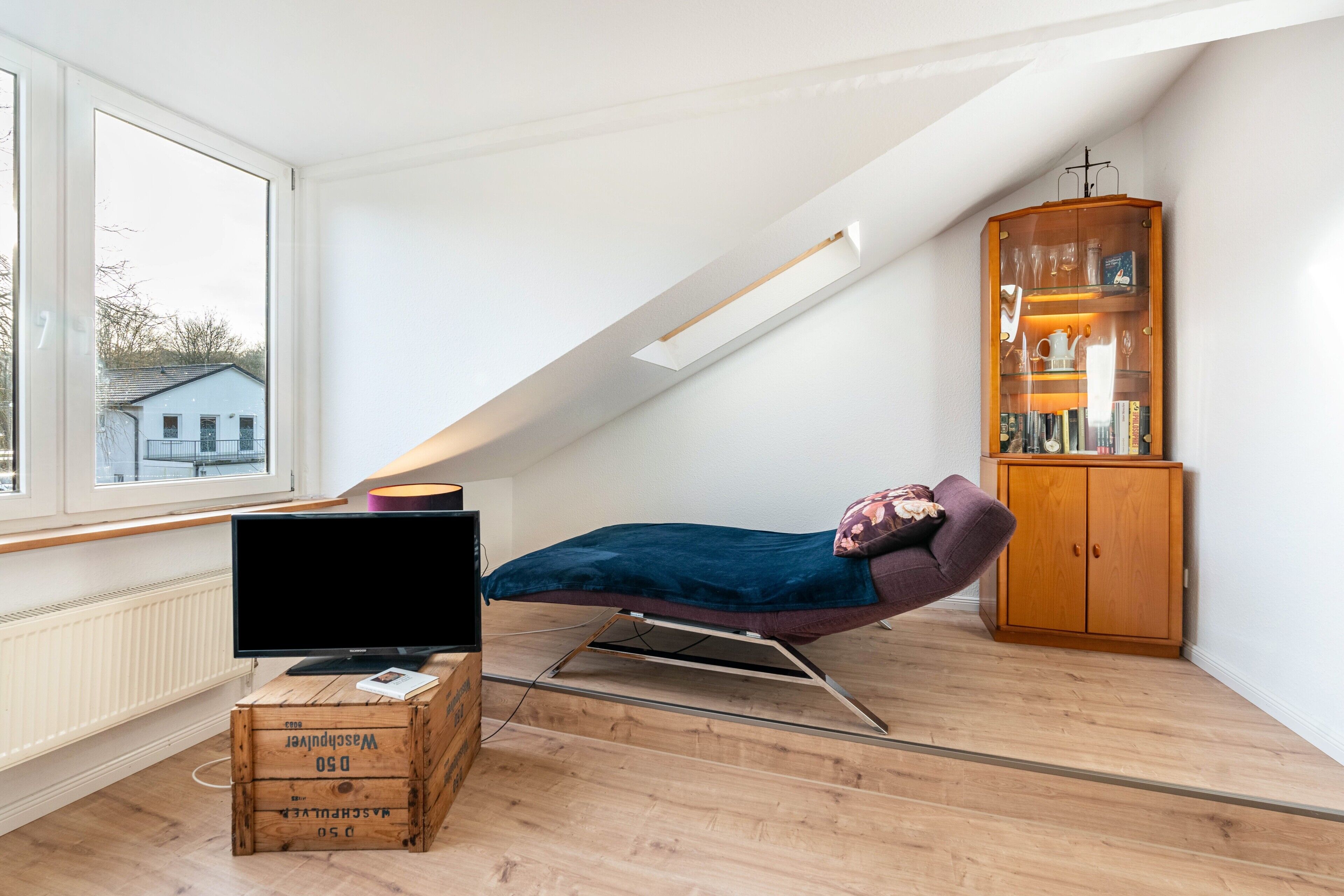 A loft bedroom with a slanted ceiling, a bed, a television on a wooden crate, a wooden cabinet, and a window with a view of a house.