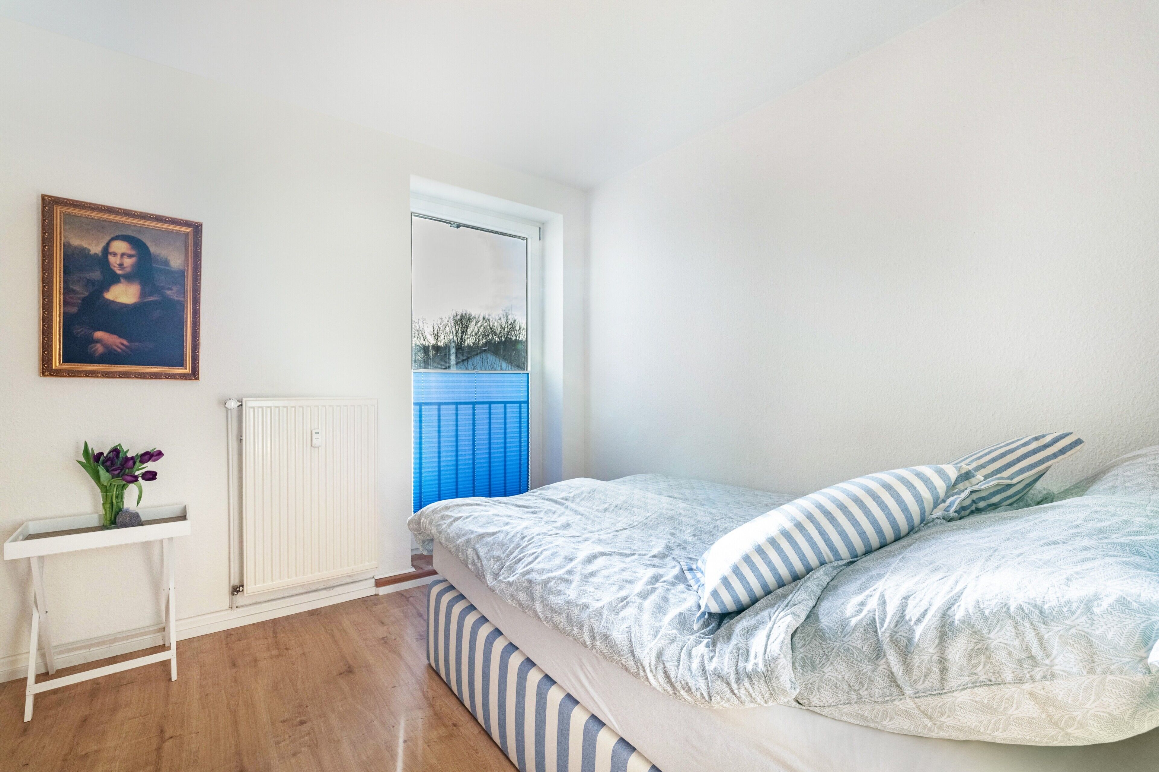 A bedroom with a bed, a bedside table with a vase, a radiator, and a window with a view.