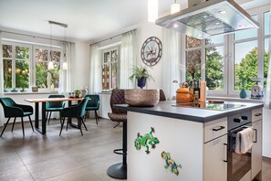 Una cocina moderna con una isla central, zona de comedor con sillas y ventanas amplias con cortinas.