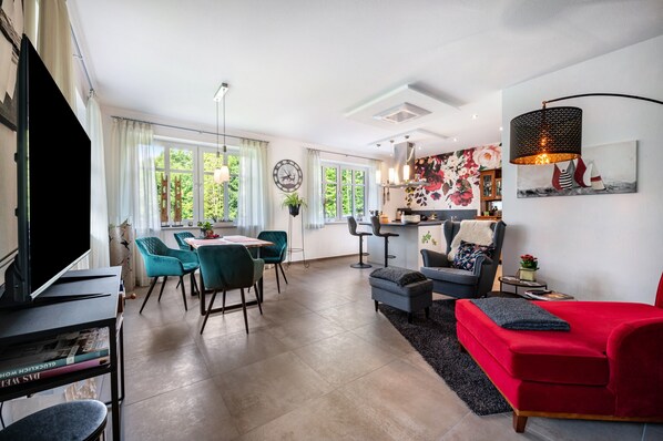 Una sala de estar moderna con un sofá rojo, sillas color turquesa, un comedor con una mesa y una cocina al fondo.