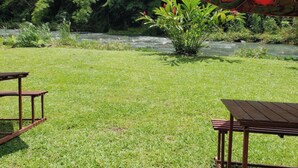 Un espacio al aire libre con césped, un río y abundante vegetación. Hay mesas de picnic y sombrillas.