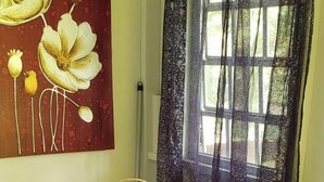Una habitación con una silla blanca, un cojín con la bandera de Puerto Rico, una mesa con una cesta y una vela, y un cuadro de flores en la pared.