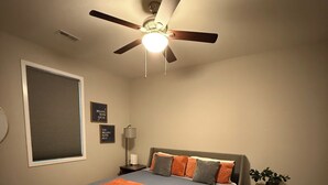 A bedroom with a bed, a ceiling fan, a nightstand, a lamp, and a window with blinds. - Fisher House Spacious Home in Tiny Home Village (Sedalia)