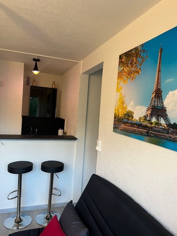 Una sala moderna con un sofá negro, dos taburetes negros, una cocina con mostrador negro y un cuadro grande de la Torre Eiffel.