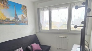 A living room with a black sofa, a small table, and a radiator. There is a window with curtains and a painting of the Eiffel Tower. - Le Cocoon / Free parking / Wifi (Belfort)