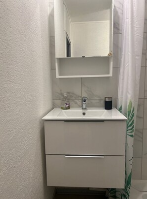 Un baño con un lavabo blanco, un espejo encima y una cortina de ducha con estampado de hojas verdes.