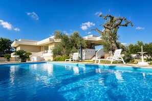 Una piscina con sillones de descanso, una casa moderna de techo plano y un árbol con ramas retorcidas.