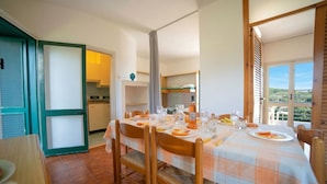 A dining area with a table set for four, chairs, a kitchen in the background, and a view through a window.