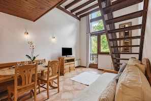 A room with a wooden dining table, chairs, a TV, a sofa, and a balcony with a wooden railing.