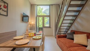 Un comedor con mesa de madera dispuesta para cuatro personas, un sofá con cojines, un televisor montado en la pared y una ventana con vistas a zonas verdes.