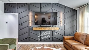 Een moderne woonkamer met een grote flatscreen televisie aan een wand met een geometrisch patroon, een bruine leren bank, een groene fauteuil en een houten salontafel.