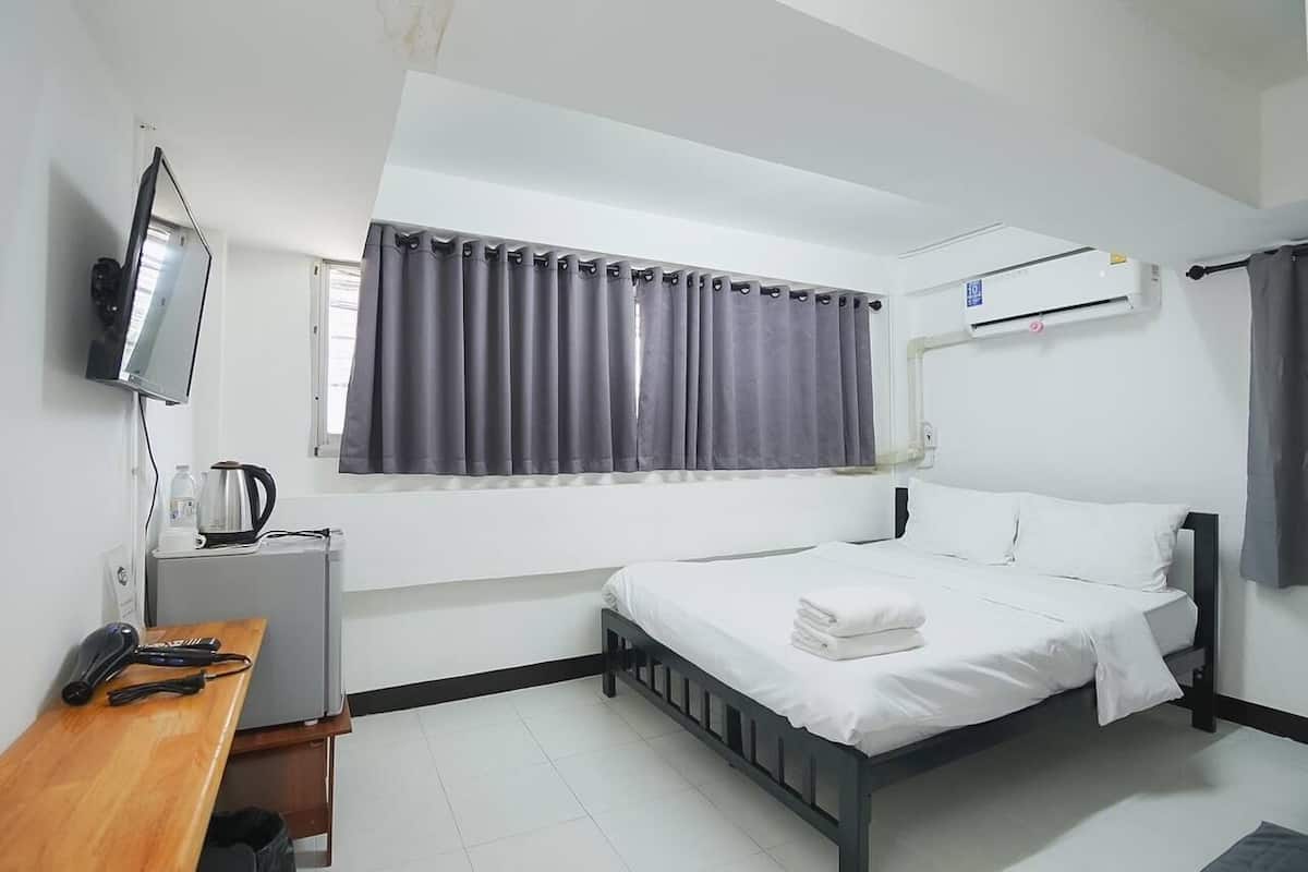 A hotel room with a bed, a desk with a kettle, a television, and an air conditioning unit.